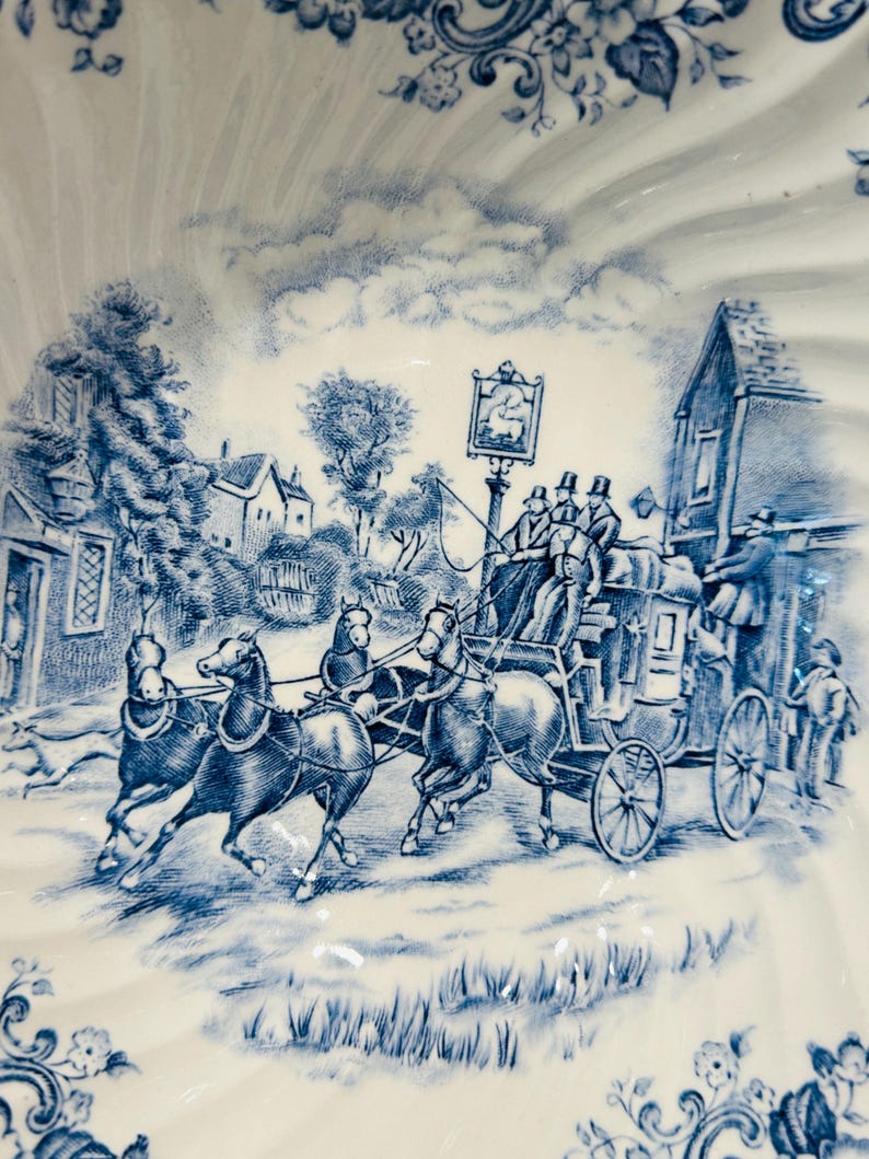 May include: A white ceramic bowl featuring a blue transferware pattern depicting a horse-drawn stagecoach traveling past a village building with a sign, surrounded by floral borders and a scalloped edge design on the rim of the dish.