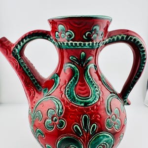 Könnte beinhalten: Roter Keramikkrug mit glänzendem Finish, verziert mit grünen Blumen- und Rankenmustern. Er hat zwei Henkel und einen Ausguss, mit einem dekorativen Rand. Das Design ist aufwendig und vintage, erinnert an traditionelle Keramik.