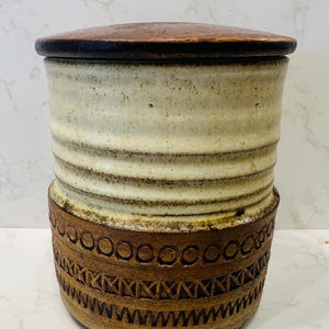 May include: A white ceramic jar with a brown wooden lid. The jar has a textured, wavy design and a brown band with a geometric pattern around the bottom.