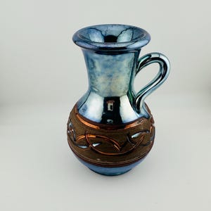 以下が含まれることがあります： 首と底にメタリックブルーの虹色の釉薬が施された陶器製のピッチャー。中央には質感のある幅広の茶色の帯があり、その帯には銅色の渦巻き模様が浮き彫りになっています。取っ手が一つ付いています。