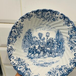 May include: A white ceramic bowl with a scalloped edge and a blue transferware pattern. The center features a detailed scene of a horse-drawn stagecoach traveling past buildings, while the rim is decorated with intricate floral motifs.