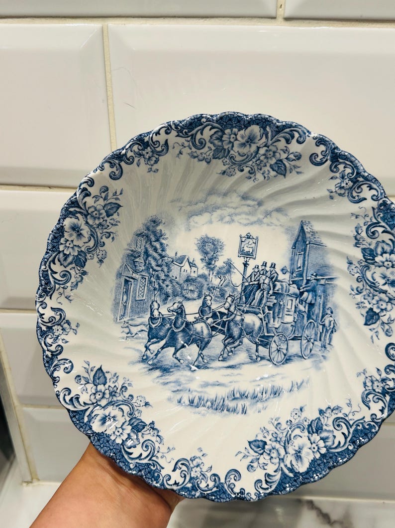 May include: A white ceramic bowl with a scalloped edge and a blue transferware pattern. The center features a detailed scene of a horse-drawn stagecoach traveling past buildings, while the rim is decorated with intricate floral motifs.
