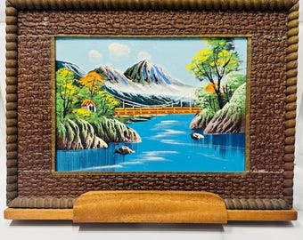 Könnte beinhalten: Gerahmtes Landschaftsgemälde, das eine Berglandschaft mit Fluss, Brücke und kleinem Haus zeigt. Das Gemälde befindet sich in einem Holzrahmen mit strukturiertem Rand und einem Holzständer. Das Kunstwerk weist blaue, grüne, gelbe und braune Farben auf.
