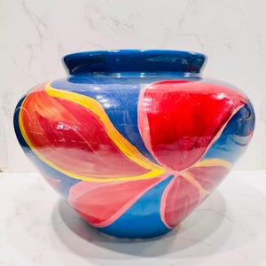 May include: A hand-painted ceramic vase with a wide body and a narrow, dark blue rim. The vase is primarily blue with large, abstract floral designs in red, pink, and yellow. The vase is a decorative item.