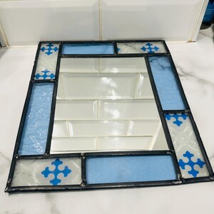 K&ouml;nnte beinhalten: Ein rechteckiger Spiegel, der mit Buntglas-Paneelen in Hellblau und Wei&szlig; gerahmt ist. Der Glasrand zeigt ein sich wiederholendes blaues kreuzartiges Muster auf strukturierten wei&szlig;en Abschnitten und hellblauen Segmenten, verbunden durch dunkles Metall.