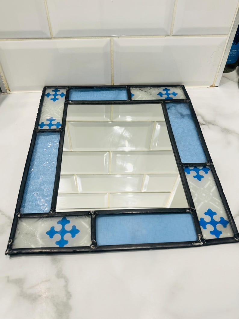 K&ouml;nnte beinhalten: Ein rechteckiger Spiegel, der mit Buntglas-Paneelen in Hellblau und Wei&szlig; gerahmt ist. Der Glasrand zeigt ein sich wiederholendes blaues kreuzartiges Muster auf strukturierten wei&szlig;en Abschnitten und hellblauen Segmenten, verbunden durch dunkles Metall.