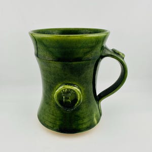 May include: A forest green ceramic mug with a glossy finish, featuring a flared rim and a curved handle. A circular medallion is embossed on the front, displaying a stylised crown symbol above the letter B, with subtle horizontal texture lines.