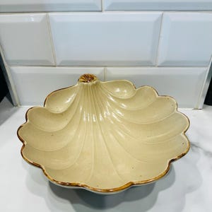 Op de afbeelding: Een beige keramische schelpvormige kom met een geschulpte rand en een bruine rand. De kom heeft een textuur met een waaiervormig patroon. De kom staat op een wit oppervlak.