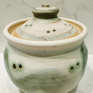 Puede incluir: Un tarro de cerámica blanco con tapa, con un esmalte verde y marrón con un patrón moteado. El tarro tiene una forma redondeada y una abertura ancha.