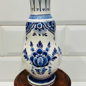 May include: A white and blue ceramic vase with a floral design, on a small, round wooden stand. The vase has a bulbous body and flared rim, with intricate blue patterns. The stand is approximately 10 cm in diameter.