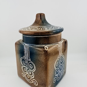 May include: A square ceramic jar with a matching lid, featuring a gradient finish that transitions from dark blue to brown. The surface is decorated with white etched swirl patterns that extend from the top rim down the sides of the container.