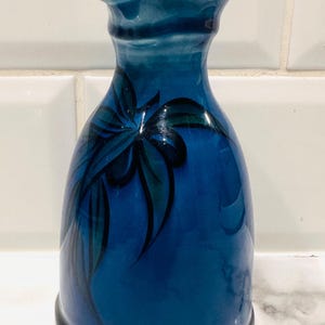 Op de afbeelding: Een blauwe keramische vaas met een uitlopende rand en een afgeronde basis. De vaas heeft een handgeschilderd ontwerp van gestileerde bladeren in blauw- en groentinten. De vaas is ongeveer 18 cm hoog.