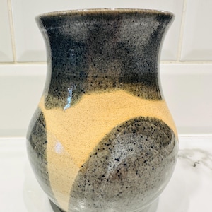May include: A handcrafted ceramic vase with a unique design. The vase features a speckled gray and black upper portion, transitioning to a tan and cream-colored lower section. The vase has a rounded body and a flared rim, perfect for displaying flowers or as a decorative piece.