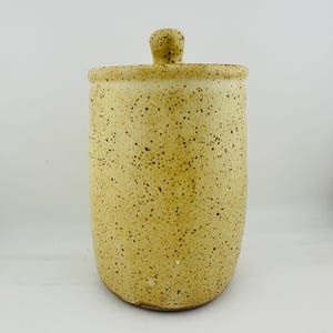 May include: A cylindrical ceramic canister with a speckled cream and yellow glaze. The container features a matching lid with a small, rounded knob handle at the top. The surface has a textured appearance with numerous dark brown speckles.