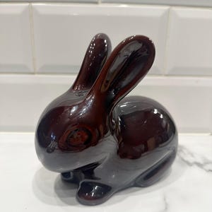 Może przedstawiać: Błyszcząca, ciemnobrązowa ceramiczna figurka królika. Królik siedzi z uniesionymi uszami. Figurka ma gładką, odblaskową powierzchnię i jest przedmiotem dekoracyjnym. Królik to klasyczny element dekoracji wielkanocnej lub wiosennej.