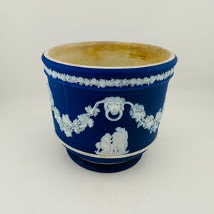 Puede incluir: Una maceta de cerámica azul profundo con adornos en relieve de color blanco. El exterior muestra una banda de motivos florales en la parte superior, asas de anillo con cabeza de león y figuras clásicas entre vides. El interior es beige.