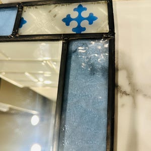 K&ouml;nnte beinhalten: Ein Buntglasspiegel mit einem dunklen Metallrahmen. Der obere Rand zeigt eine wei&szlig;e Glasscheibe mit einem blauen kreuzf&ouml;rmigen Muster. Ein vertikales Paneel aus strukturiertem, hellblauem Glas verl&auml;uft entlang der Seite neben der Spiegelfl&auml;che.