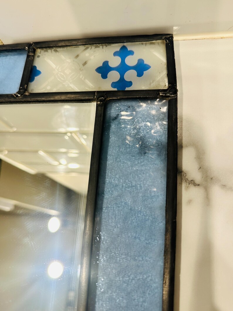 K&ouml;nnte beinhalten: Ein Buntglasspiegel mit einem dunklen Metallrahmen. Der obere Rand zeigt eine wei&szlig;e Glasscheibe mit einem blauen kreuzf&ouml;rmigen Muster. Ein vertikales Paneel aus strukturiertem, hellblauem Glas verl&auml;uft entlang der Seite neben der Spiegelfl&auml;che.