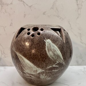May include: A brown ceramic vase with a spherical form and a series of holes at the top. The vase features a relief design of two birds in white. The vase is a decorative item.