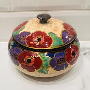 Può includere: Un barattolo rotondo in ceramica con coperchio, beige. Il barattolo è decorato con fiori di papavero rossi e viola con foglie verdi e dettagli dorati. Il coperchio ha un piccolo pomello marrone scuro.