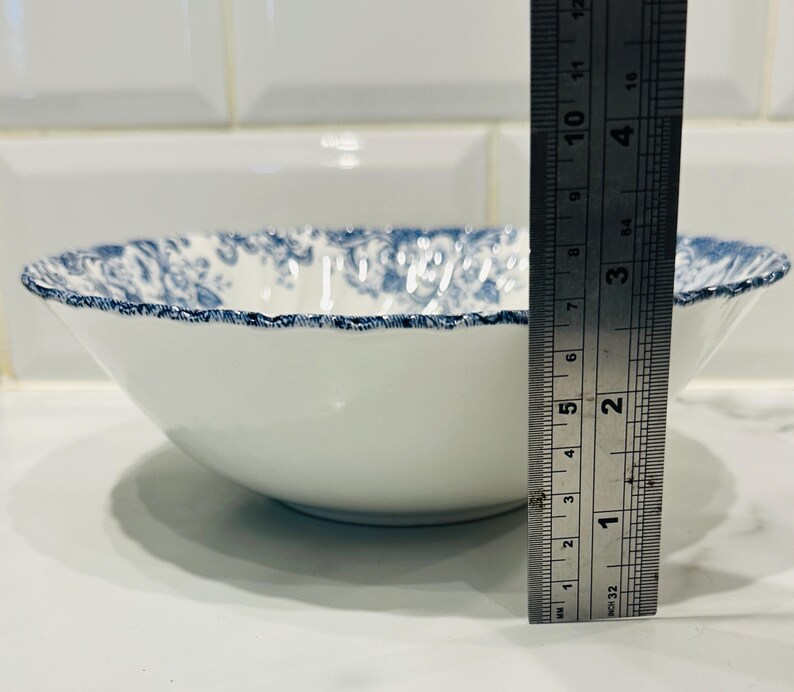 May include: A white ceramic bowl with a blue floral pattern along the scalloped rim. A metal ruler is positioned vertically against the side of the bowl, showing a height of approximately 2.5 inches from the base to the top edge of the rim.