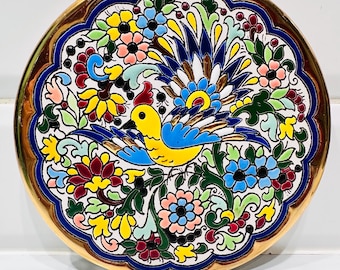 Könnte beinhalten: Ein runder, dekorativer Keramikteller mit goldenem Rand und einem lebendigen Blumen- und Vogeldesign. Der Teller zeigt einen zentralen Vogel mit blauen Flügeln und gelbem Körper, umgeben von bunten Blumen in Rosa, Gelb und Blau. Der Teller hat einen Durchmesser von etwa 20 cm.