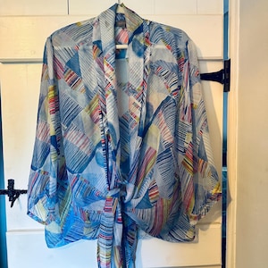 May include: A lightweight, sheer kimono-style cardigan featuring a geometric pattern in shades of blue, white, yellow, and red. The garment has wide, three-quarter length sleeves, an open front, and a tie-front closure at the bottom hem.