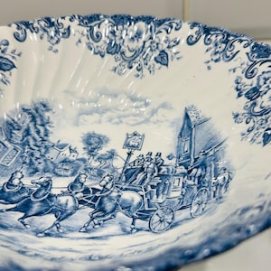 May include: A white ceramic bowl with a scalloped edge and a blue transferware pattern. The design features a horse-drawn stagecoach traveling past a building with a sign, surrounded by floral motifs along the rim and a cloudy sky background.