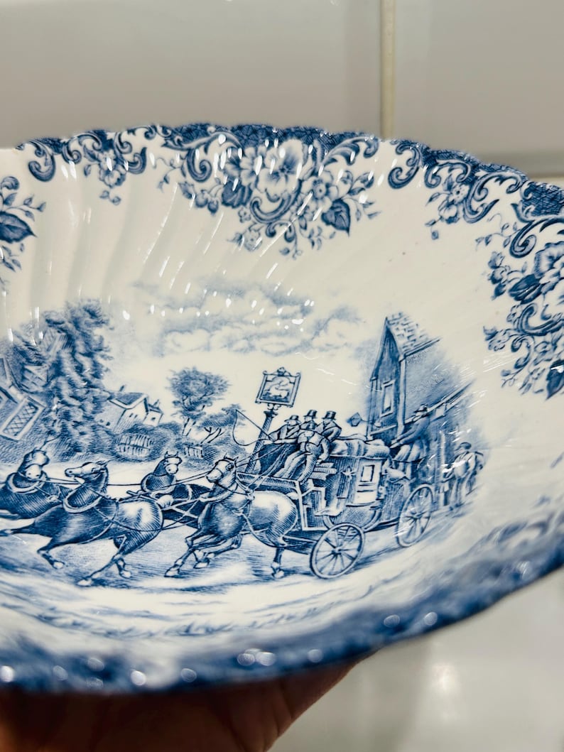 May include: A white ceramic bowl with a scalloped edge and a blue transferware pattern. The design features a horse-drawn stagecoach traveling past a building with a sign, surrounded by floral motifs along the rim and a cloudy sky background.