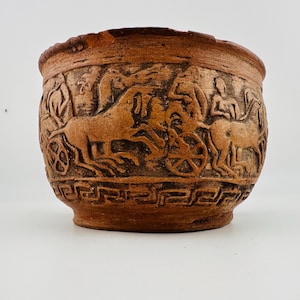 May include: A terracotta-colored bowl featuring a relief carving of figures riding in horse-drawn chariots around the exterior. The base of the bowl is decorated with a repeating geometric Greek key pattern in a dark brown finish.