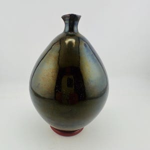 Könnte beinhalten: Eine birnenförmige Keramikvase mit einer glänzenden, dunkelmetallischen Glasur, die zwischen tiefem Bronze, Schwarz und subtilen Blautönen wechselt. Die Oberfläche hat ein glattes, reflektierendes Finish mit einem schmalen Hals und einem roten Fuß.