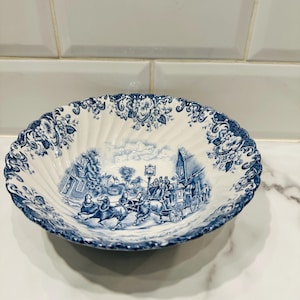 May include: A round, scalloped-edge ceramic bowl featuring a blue and white transferware pattern. The center depicts a horse-drawn carriage scene with figures, while the rim is decorated with a detailed floral motif in matching blue ink.
