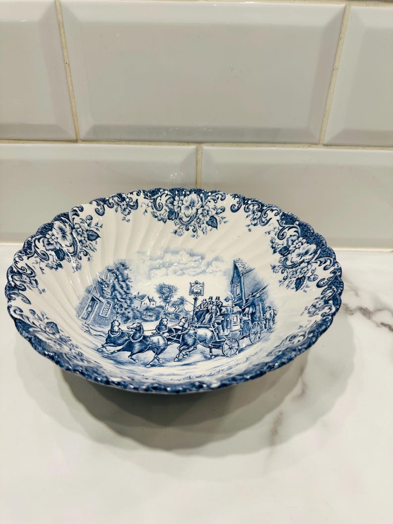 May include: A round, scalloped-edge ceramic bowl featuring a blue and white transferware pattern. The center depicts a horse-drawn carriage scene with figures, while the rim is decorated with a detailed floral motif in matching blue ink.