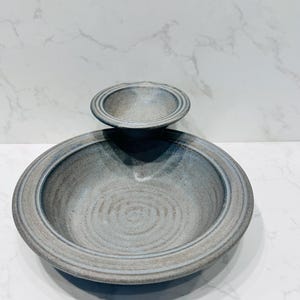 May include: Two-piece ceramic serving bowl set. The larger bowl is grey with a wide rim and a circular design in the centre. A smaller bowl sits on top. The bowls have a rustic, handmade appearance.