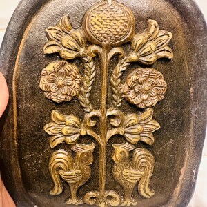 May include: A dark, oval-shaped decorative plaque with a raised, gold-colored floral and bird design. The design includes a central stem with flowers, a pomegranate, and two rooster-like birds at the base. The plaque has a textured, aged appearance.
