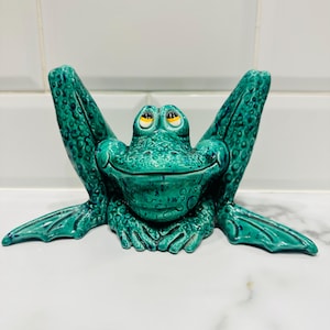 May include: A teal ceramic frog ornament. The frog has large, yellow-eyed, smiling face and outstretched arms. The textured surface adds detail to the design. The frog is a decorative item.