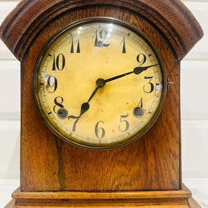 May include: Antique wooden mantel clock with a round face. The clock has a light yellow face with black numerals and hands. The clock is housed in a dark brown wooden case with an arched top and a rectangular base. The clock face is protected by a glass cover.