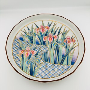 May include: A ceramic plate with a floral design. The plate features pink and blue irises, green leaves, and a blue and white checkered pattern. It has a white rim and a brown edge. The design is set against a white background.