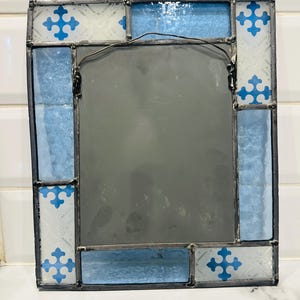 K&ouml;nnte beinhalten: Ein rechteckiger Spiegel mit einem Rahmen aus Buntglas und dunklen Metalll&ouml;tstellen. Der Rand besteht aus abwechselnden Paneelen aus strukturiertem wei&szlig;em Glas mit blauen Kreuzmustern und einfarbigem hellblauem Glas. Ein Drahtaufh&auml;nger ist hinten.