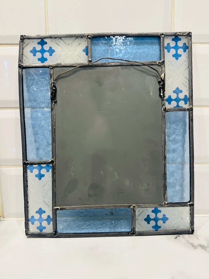 K&ouml;nnte beinhalten: Ein rechteckiger Spiegel mit einem Rahmen aus Buntglas und dunklen Metalll&ouml;tstellen. Der Rand besteht aus abwechselnden Paneelen aus strukturiertem wei&szlig;em Glas mit blauen Kreuzmustern und einfarbigem hellblauem Glas. Ein Drahtaufh&auml;nger ist hinten.