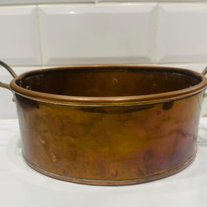May include: A round copper pot with a polished metallic finish and two curved metal handles on opposite sides. The vessel features a wide, open top and a smooth, cylindrical body with visible variations in the warm, reddish-brown copper tone.