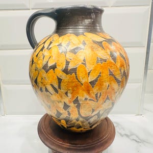 Puede incluir: Una jarra de cerámica con borde y asa marrón oscuro, adornada con un patrón de diseños de hojas naranjas y amarillas. La jarra se encuentra sobre un pequeño soporte redondo de madera.