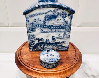 Könnte beinhalten: Eine blau-weiße Porzellan-Teedose und eine kleine runde Schmuckschatulle mit Landschaftsdesign. Die Teedose ist hausförmig und steht auf einem kleinen Holzständer. Die Schmuckschatulle ist rund mit einer passenden Landschaftsszene.
