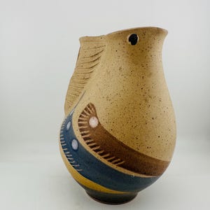 May include: A speckled beige ceramic pitcher with a bird-like silhouette. The body features a curved handle, a small black eye detail, and decorative bands of blue, brown, and yellow paint with white dots along the lower section of the vessel.