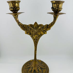 May include: A gold-toned metal candelabra featuring two candle holders with fluted rims. The central stem and base are decorated with embossed floral and leaf motifs. The structure has a symmetrical design with a wide, circular, textured base.