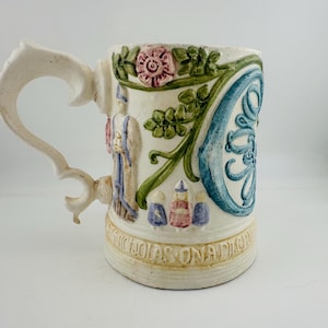 Könnte beinhalten: Ein cremefarbener Keramikkrug mit erhabenen, handbemalten Reliefs einer rosa Rose, grünem Blattwerk, einem blauen Wirbelmuster und kleinen Figuren. Am Boden befindet sich der geprägte Text "ST. NICHOLAS ON A PILGRIMAGE".