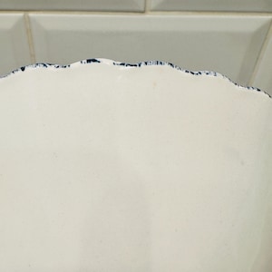 May include: A cream-colored ceramic bowl featuring a scalloped edge rim. The rim is decorated with a dark blue speckled or sponged pattern that follows the wavy contour of the bowl's top border, contrasting against the smooth, solid surface.
