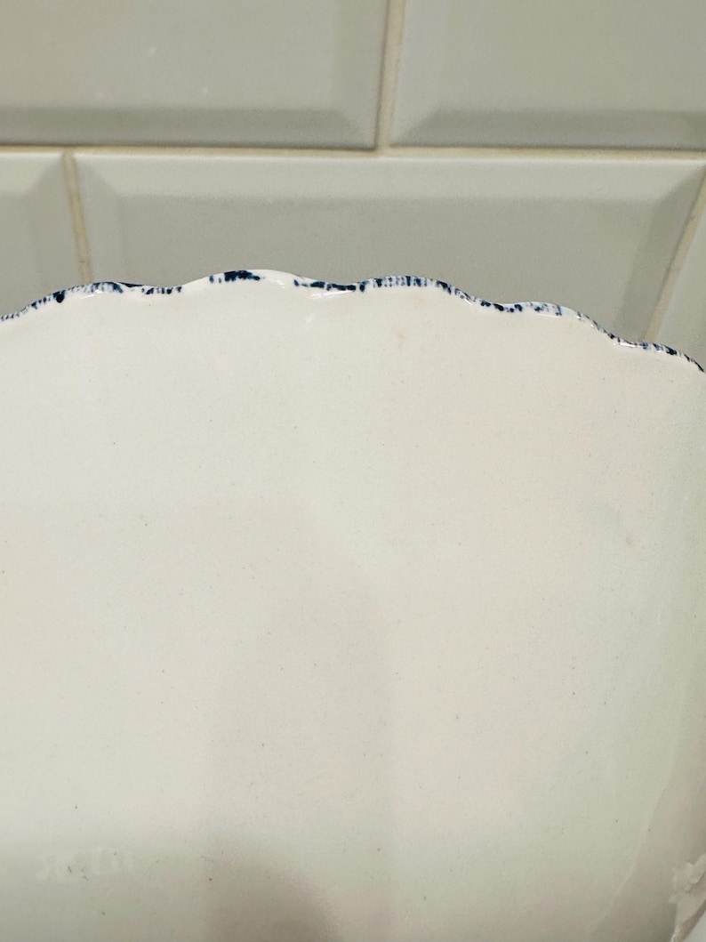 May include: A cream-colored ceramic bowl featuring a scalloped edge rim. The rim is decorated with a dark blue speckled or sponged pattern that follows the wavy contour of the bowl's top border, contrasting against the smooth, solid surface.