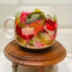 May include: A white ceramic mug with a colourful abstract design in red, pink, green, and orange. The mug is decorated with gold accents and sits on a small, round, wooden stand with four legs.