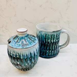 May include: A ceramic mug and sugar bowl set. The mug has a handle and a textured, teal and black glaze. The sugar bowl has a lid with a small spoon notch. The set is a unique addition to any kitchen.